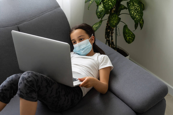 Smart Little Girl wearing mask Does Homework in Her Living Room. Shes Sitting and Uses Laptop. - Foto, afbeelding