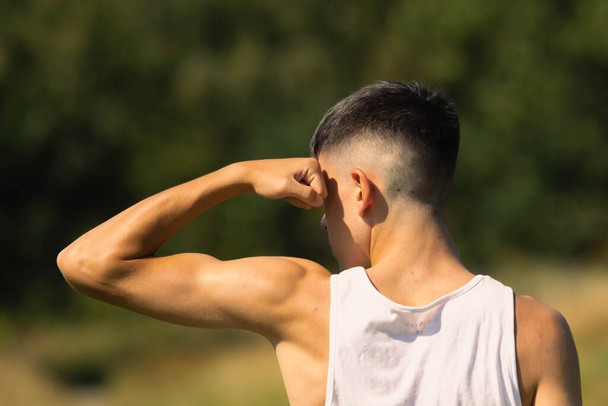 An 18 year old teen boy in a sleeveless top on a late summer's day, flexing his arm muscles - 写真・画像