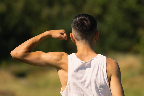 An 18 year old teen boy in a sleeveless top on a late summer's day, flexing his arm muscles - Photo, Image