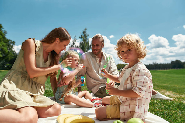 Loving parents spending time with two little kids, blowing soap bubbles in the park. Family having picnic in nature on a summer day - Foto, imagen