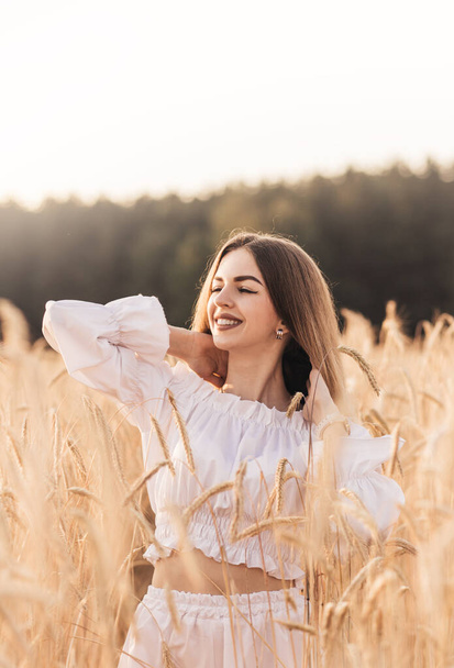 A young beautiful woman walks through a wheat field in white clothes - Фото, изображение