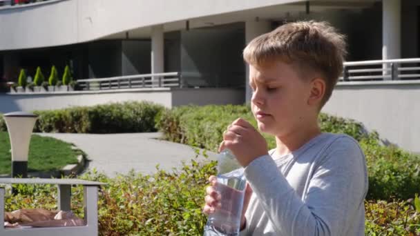 Schoolboy sentado no banco e bebe água tranquila de garrafa de plástico. - Filmagem, Vídeo