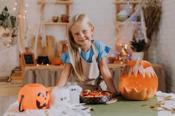 Blonde baby girl with long hair in an apron in the kitchen, decorated with pumpkins and garlands for Halloween, prepares a focaccia pie. space for text. High quality photo - Fotó, kép
