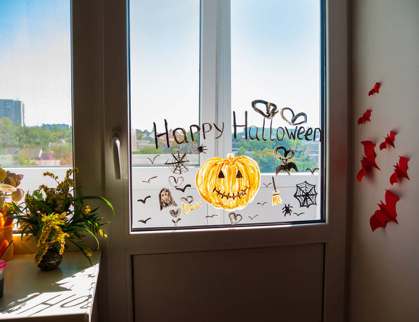 Pencereye balkabağı çizen çocuk Cadılar Bayramı 'nı kutluyor. Küçük çocuk odanın içini kağıt yarasalarla süslüyor. Yaratıcı aile eğlencesi, yeni gerçeklik, güz tatilini kutluyor. - Fotoğraf, Görsel