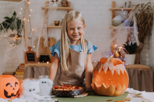 Bambina bionda dai capelli lunghi in grembiule in cucina, decorata con zucche e ghirlande per Halloween, prepara una focaccia. spazio per il testo. Foto di alta qualità - Foto, immagini