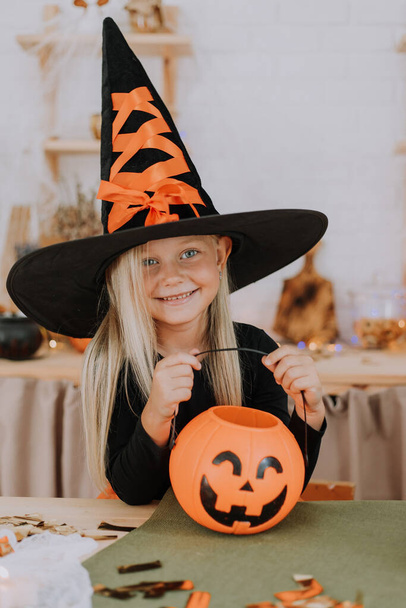 Portret van een klein blond meisje met lang haar in een heksenkostuum met een enorme heksenhoed, die een pompoenvormige snoepmand in haar handen houdt. Halloween concept. Ruimte voor tekst. Hoge kwaliteit foto - Foto, afbeelding