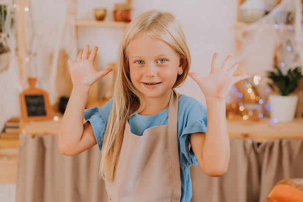 Blondes kleines Mädchen mit langen Haaren in einer Schürze in der Küche, dekoriert mit Kürbissen und Girlanden zu Halloween, bereitet eine Focaccia-Torte zu. Raum für Text. Hochwertiges Foto - Foto, Bild