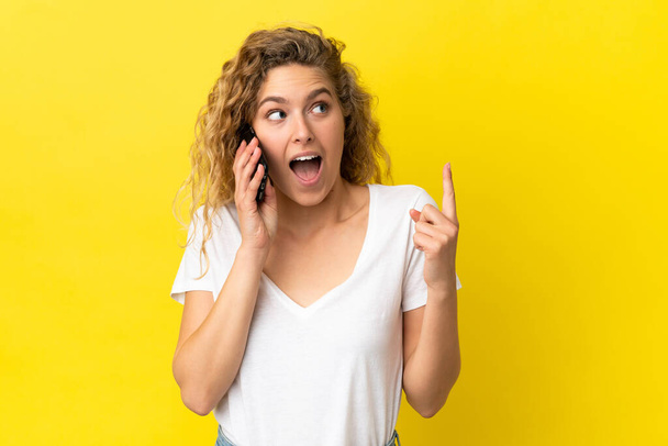 Nuori blondi nainen käyttää matkapuhelinta eristetty keltaisella taustalla ajattelu ajatus sormella ylöspäin - Valokuva, kuva