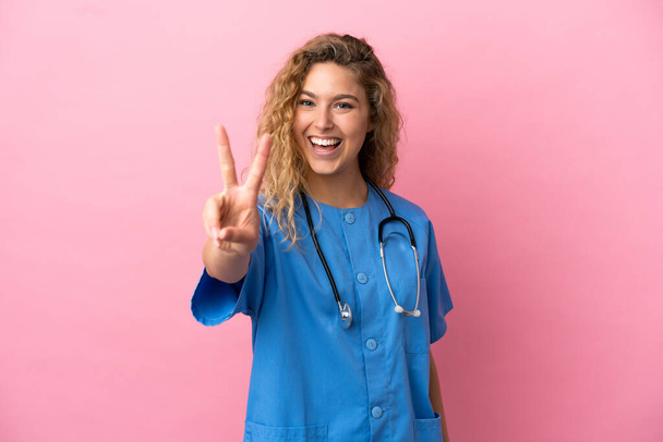 Young surgeon doctor woman isolated on pink background smiling and showing victory sign - Foto, afbeelding
