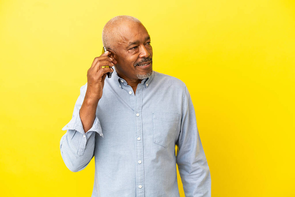 Kubanischer Senior isoliert auf gelbem Hintergrund und hält ein Gespräch mit dem Handy mit jemandem - Foto, Bild