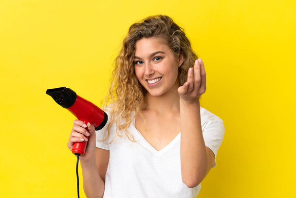 Young blonde woman holding a hairdryer isolated on yellow background making money gesture - Photo, Image