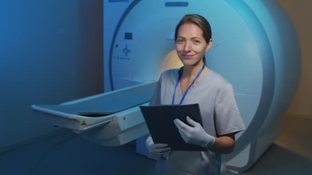 現代の診療所でMRI装置のスキャナに対してポーズをとる笑顔女性放射線技師のスローモ中肖像 - 映像、動画