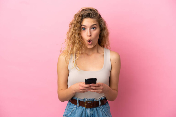Mujer rubia joven aislada sobre fondo rosa mirando a la cámara mientras usa el móvil con expresión sorprendida - Foto, Imagen