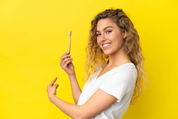 Νεαρή καυκάσια γυναίκα που κρατάει ένα βουρτσισμένο δόντι απομονωμένο σε κίτρινο φόντο που δείχνει προς τα πίσω - Φωτογραφία, εικόνα
