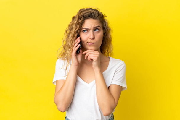 Nuori blondi nainen käyttää matkapuhelinta eristetty keltaisella taustalla, jolla on epäilyksiä ja ajattelua - Valokuva, kuva