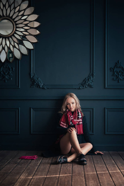Beautiful blonde woman in a blue autumn coat and a red scarf. Bare legs in contrast. Is sitting. Autumn or winter indoor portrait. - Zdjęcie, obraz