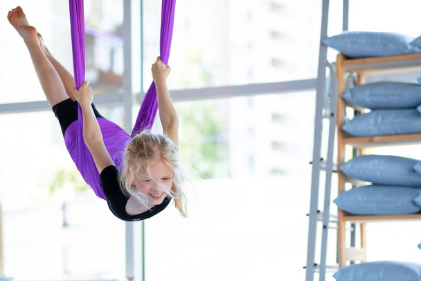 young smiling girl practice in aero stretching swing in purple hammock in fitness club. kids Aerial flying yoga exercises - Fotó, kép