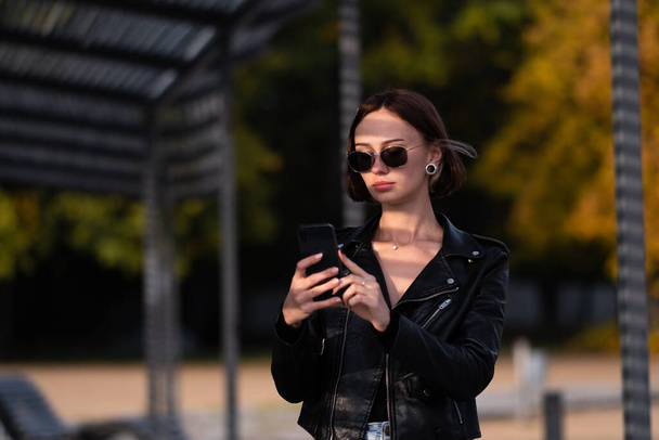 短い髪の黒い革のジャケットを着た美しい十代の少女は、電話で自撮りをします。サングラスのDevuksha通りに電話を見てください。. - 写真・画像
