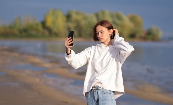 A girl in a white sweater looks at the phone on the bank of the river in autumn. Young woman walks in nature. - Photo, Image