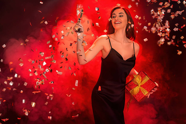 Excited woman in black dress holds the gift red box and glass of champagne. Party time. Holiday concept - Foto, Bild