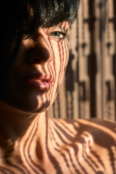 Natürliche weibliche Schönheit mit Sonnenlicht-Schatten im Gesicht und nackten Schultern mit Fokus auf das entfernte Auge in einer zugeschnittenen Nahaufnahme - Foto, Bild