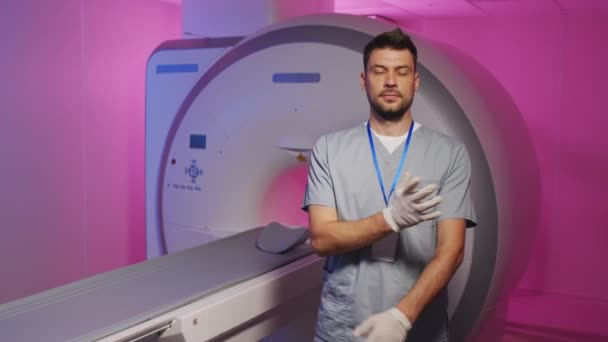 紫色の照明でMRiマシンと現代の放射線治療室に立って折り畳まれた手でポーズをとるスクラブの若い男性医師の人形入れ遅い肖像画 - 映像、動画
