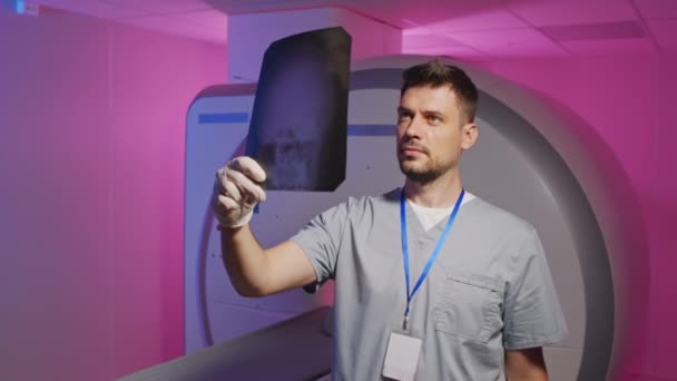 Retrato medio lento de un joven especialista en radiología o neurología masculino que mira la radiografía cerebral del paciente de pie contra la máquina de resonancia magnética en la habitación con iluminación púrpura - Imágenes, Vídeo