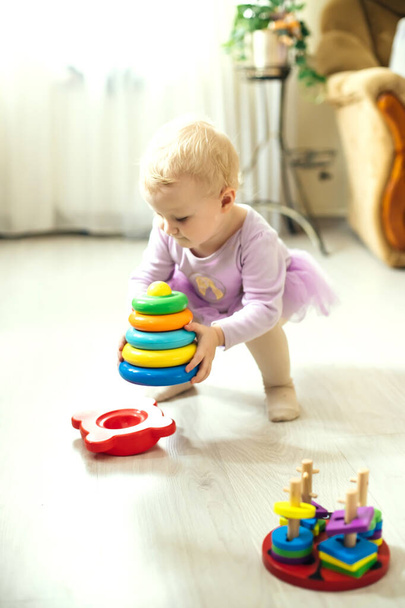 girl plays logical pyramid on floor in living room on sunny day. Montessori wooden toy folded pyramid. Circle, quadra, triangle, rectangle wooden elements of children's toys. Multi-colored toy blue, yellow, red, green - Photo, Image