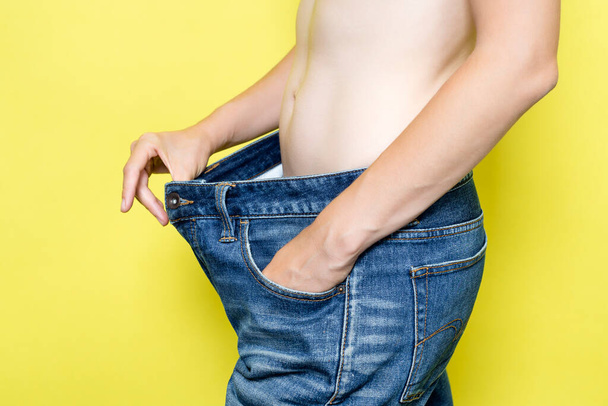 Концепция питания, правильное питание, потеря веса. Тонкая женщина, демонстрирующая джинсы и свой худый вес. Женщина в огромных джинсах изолирована на жёлтом фоне - Фото, изображение