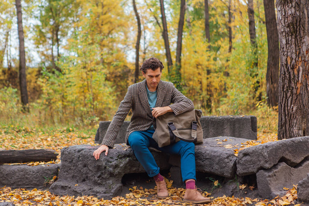 Tall handsome man dressed in a brown jacket posing with a bag on the rocks in autumn forest - Fotografie, Obrázek