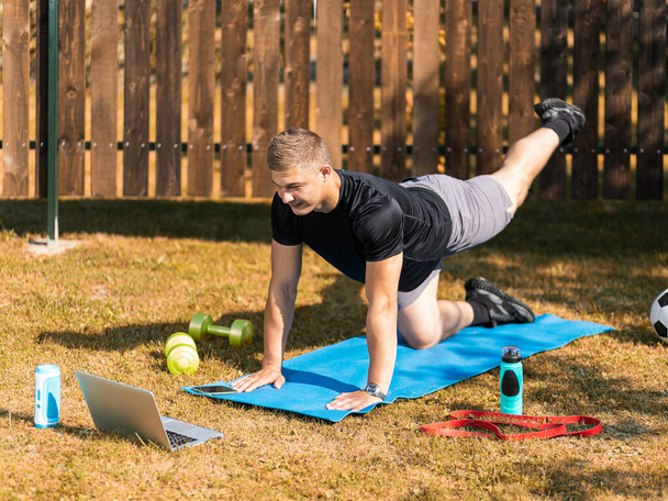 The young man goes in for sports outside. Cheerful sportsman with blonde hair makes the lunges on lawn in garden, next to there is a laptop with online training - Photo, Image