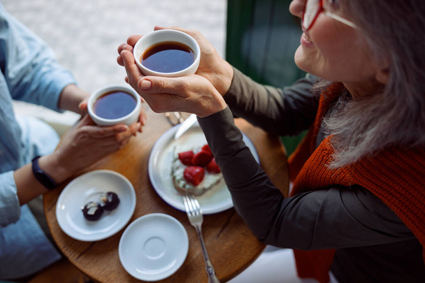 Uśmiechnięta dojrzała kobieta z przyjacielem trzymać filiżanki herbaty siedzi przy małym stole w kawiarni - Zdjęcie, obraz