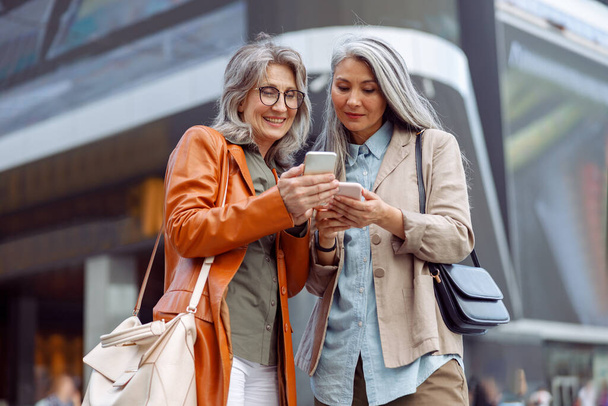 Positive grey haired ladies look at mobile phones on modern city street - Foto, Imagen