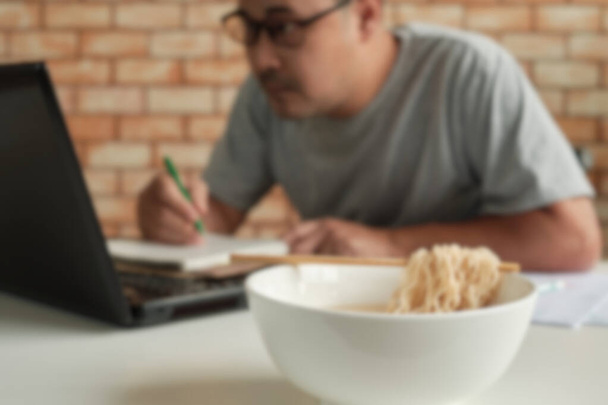 Blur tiro foco, trabalhador do sexo masculino tailandês ocupado trabalhando com laptop, usar pauzinhos para comer macarrão instantâneo durante as pausas de almoço do escritório, porque rápido e saboroso. Ao longo do tempo comida rápida asiática, estilo de vida pouco saudável. - Foto, Imagem