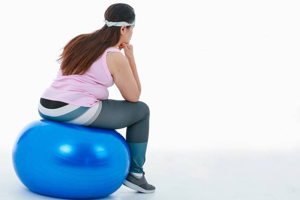 Isolated studio shot of Asian depressed stress tired big fat plus size girl in sport clothing with headband turn back sit on blue exercise fitball thinking giving up on workout on white background. - Photo, Image