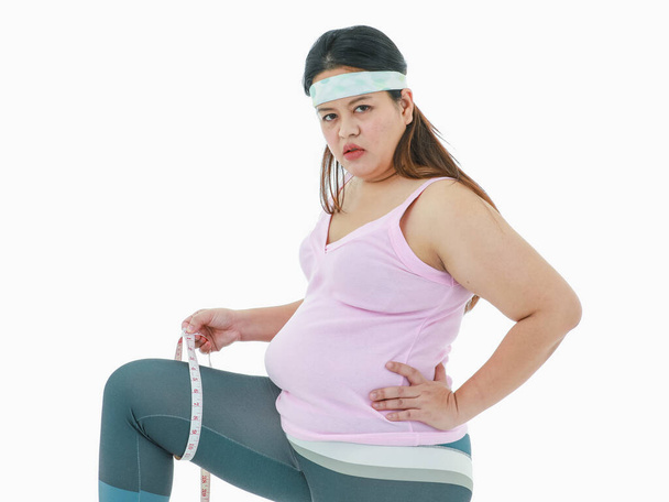 Isolated studio shot of Asian sad depressed stress worry unhealthy big fat belly stomach girl in sport clothing leggings with headband sit measuring leg with measure tape in front white background. - Photo, Image