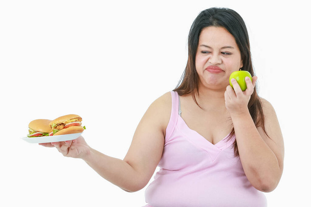 izolowane studio strzał z Azji koncentrat stres Duży tłuszcz otyłość dziewczyna zdecydować się jeść zdrowe organiczne zielone jabłko w dłoni i nienawiść nie lubi niezdrowe cheeseburgery w przednim białym tle. - Zdjęcie, obraz
