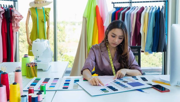 Lindo y enérgico diseñador asiático femenino que trabaja en el escritorio en la sala de diseño entre la ropa y la muestra de color de la tela. - Foto, Imagen