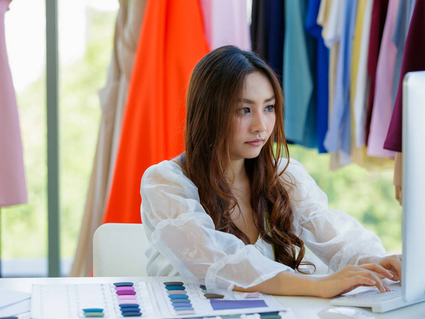 Söpö ja energinen naispuolinen aasialainen suunnittelija työskentelee työpöydällä suunnitteluhuoneessa vaatteiden ja kankaan värinäytteen joukossa. - Valokuva, kuva