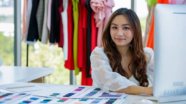 Cute i energiczny kobieta projektant Azji pracuje przy biurku w pokoju projektowania wśród ubrań i próbki kolorów tkanin. - Zdjęcie, obraz