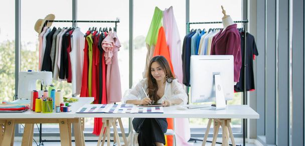 服や生地の色のサンプルの中でデザインの部屋でデスクで働く可愛くて活気に満ちた女性アジアのデザイナー. - 写真・画像