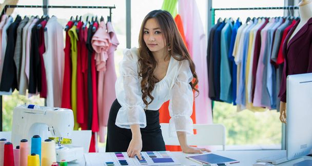 Гарненька і енергійна жінка-азіатський дизайнер працює за столом у кімнаті дизайну серед одягу і зразків тканини.. - Фото, зображення