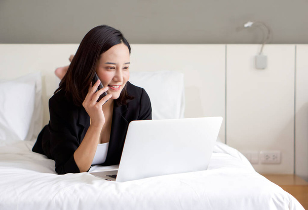 Azjatka w czarnym garniturze używa laptopa i telefonu komórkowego do pracy rano w łóżku. - Zdjęcie, obraz