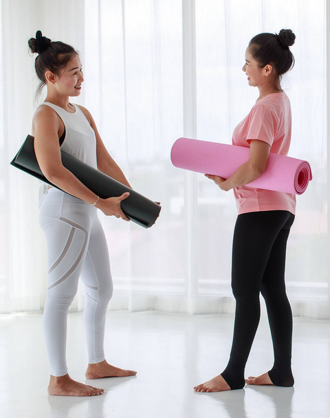 Healthy asian women in comfy sportswear holding yoga mat, enjoy talking together with happy smiling, and feel more relaxing after finish yoga training class indoors - Photo, Image