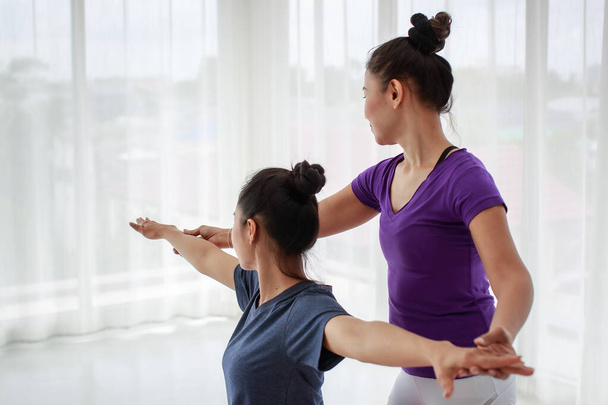 Side view portret van vrouwelijke yoga trainer staan achter de student om haar te helpen de krijger poseren en corrigeren van haar houding in fitness studio of thuis - Foto, afbeelding