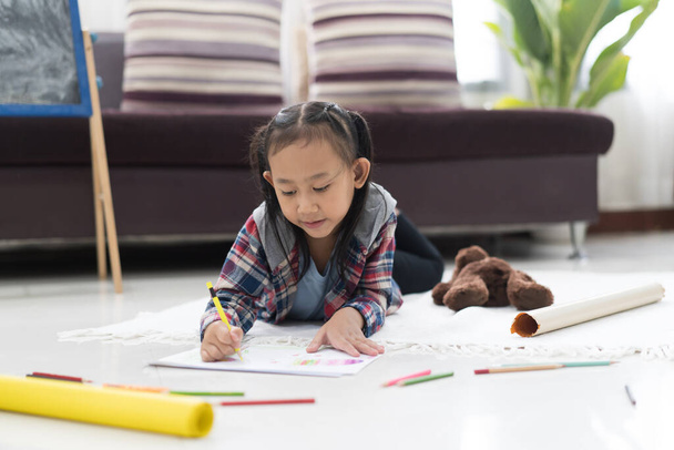 Happy cute asian little girl drawing using colorful pencils while lying on the floor at home - Foto, afbeelding