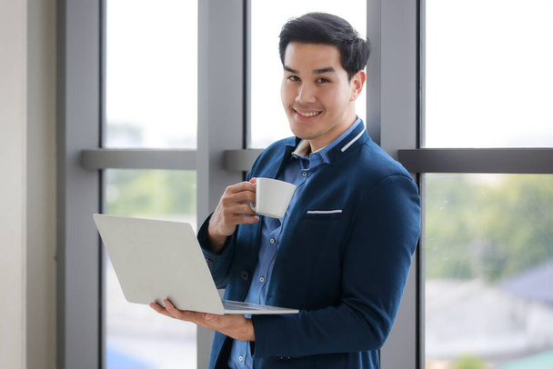 Modern takım elbiseli yakışıklı bir işadamı ofiste dizüstü bilgisayarla çalışırken bir fincan kahve içmekten zevk alır. Nazik bir gülümsemeyle kameraya bakar. - Fotoğraf, Görsel