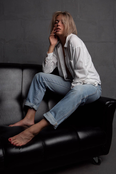 A beautiful and sexy young sensual blonde in a white shirt and jeans poses on a black sofa. Studio photo on a dark background - 写真・画像