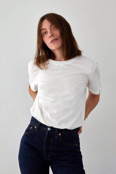 Positive female model wearing white t shirt and jeans standing with hands on waist against white background and looking at camera - Fotó, kép