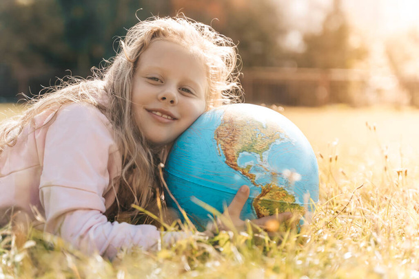 Child with Earth Globe. Portrait of a blonde European girl of elementary school with an educational globe of the planet Earth in nature in sunny weather. Study, education, conservation and - Photo, image